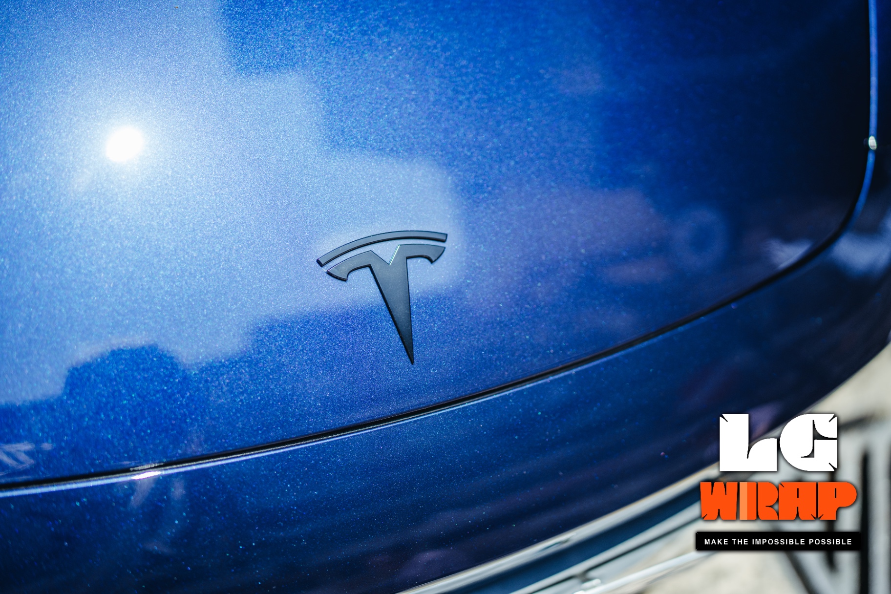 Close-up of a dark blue Tesla rear T badge, showing the deeper layering of the original pearl blue under clear PPF