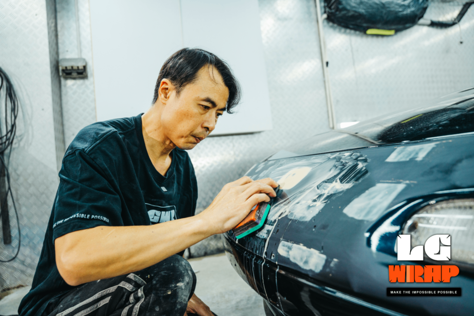 Technician crouching to apply body filler to the front bumper of a Mazda MX-5 NA