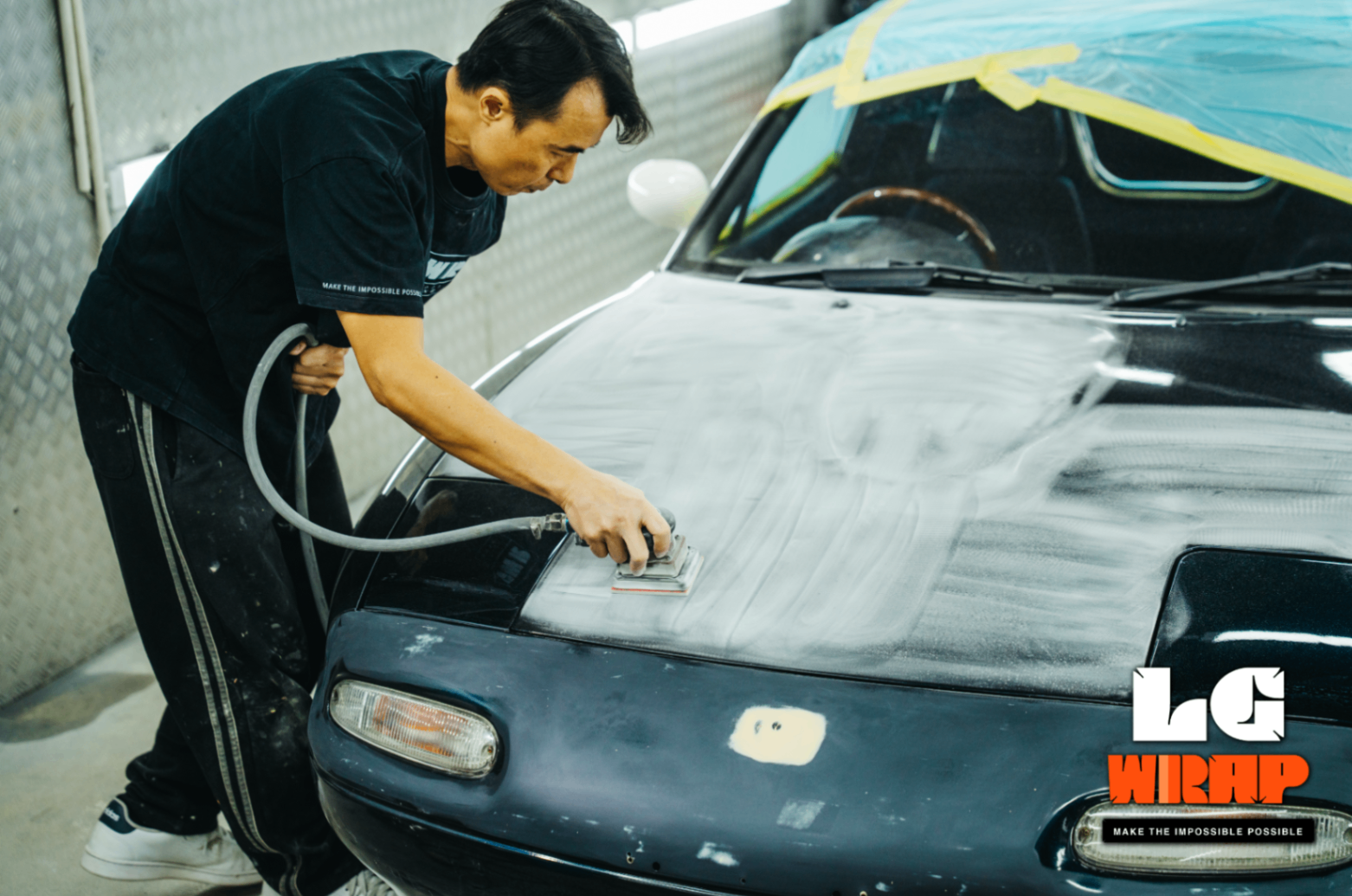 Technician performing machine sanding on the front of a Mazda MX-5 NA