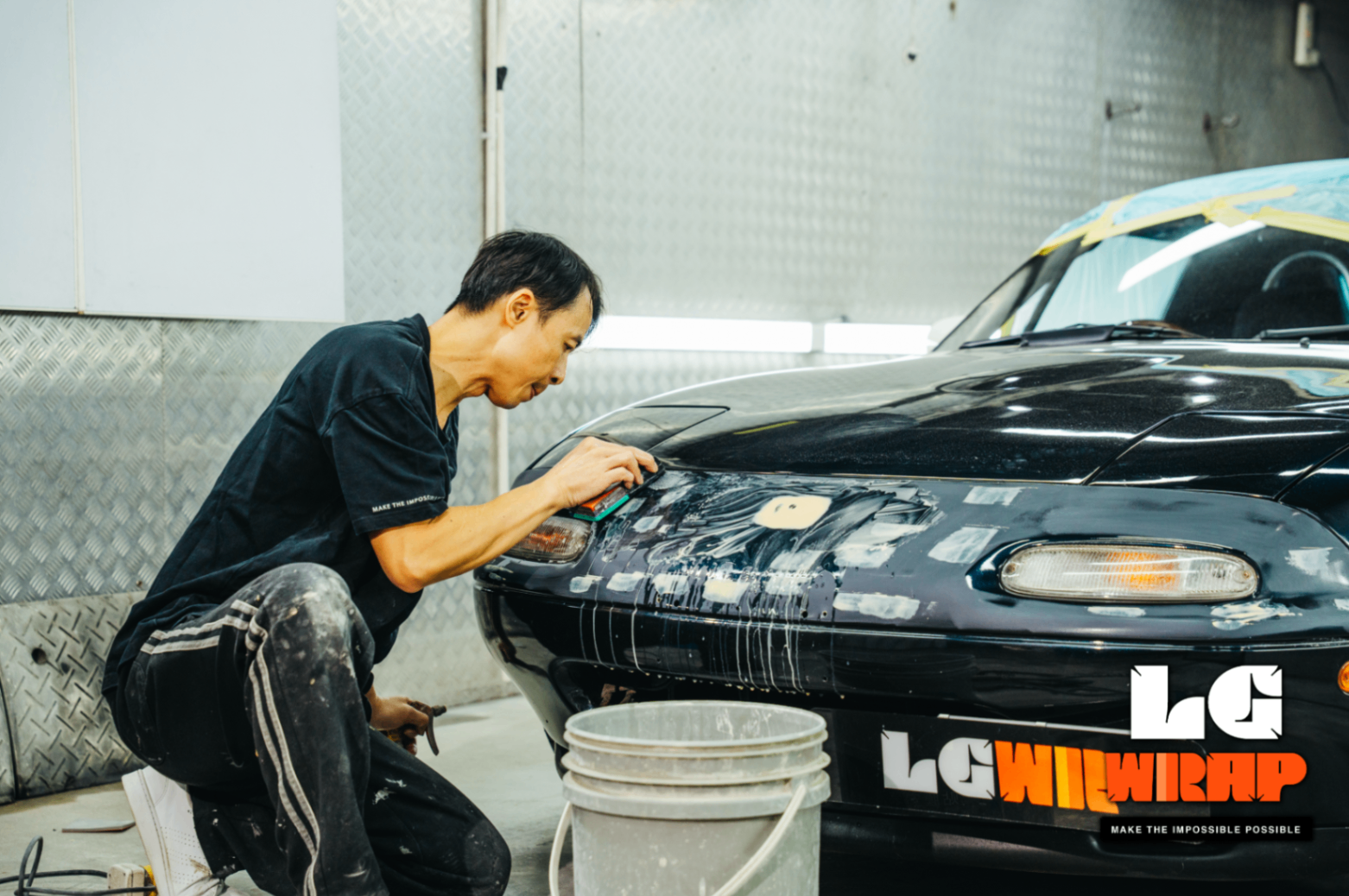 Technician performing detailed body filler repair on the front of a Mazda MX-5 NA
